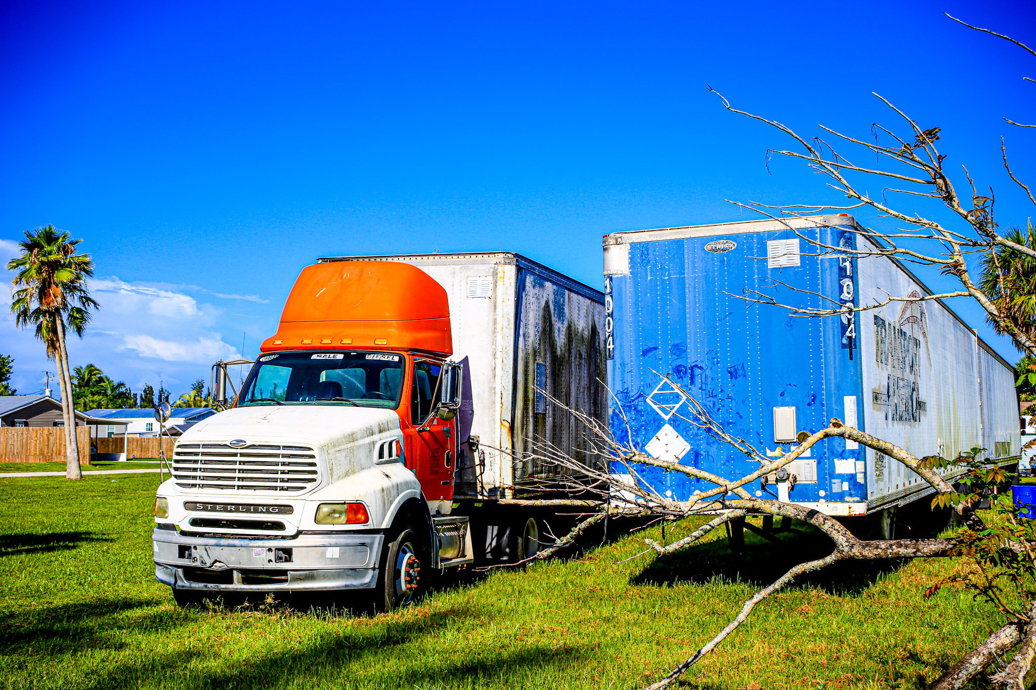 Abandoned in Time A large orange semi-truck is attached to a white trailer with blue accents, parked on a grassy area under a blue sky.